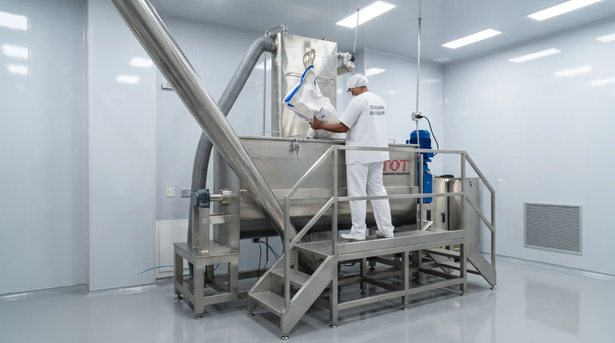 Operador en sala limpia cargando ingredientes en un Ribbon Blender de acero inoxidable fabricado por TOT, conectado a una línea de transporte neumático.